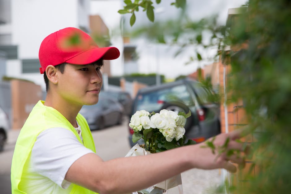 A young male florist wearing a bright red cap and a neon yellow vest arranges a bouquet of fresh white roses and hydrangeas, holding the flowers with her right hand while positioning them against a gr