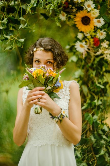 A young woman with short, curly brown hair and a delicate floral headband holds a vibrant bouquet of fresh flowers, including yellow, purple, and pink blooms with green leaves. She is dressed in a sle