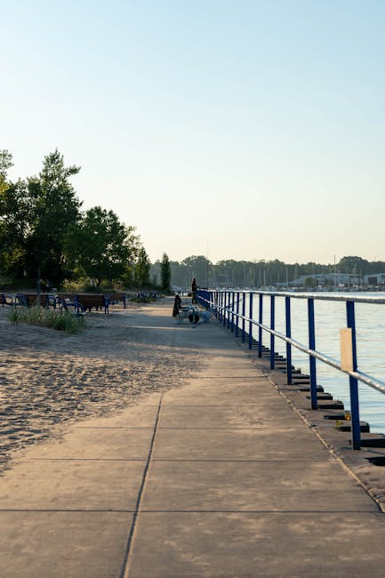 The image shows a paved promenade alongside a body of water, illuminated by soft natural light, likely during late afternoon or early evening. On the left, there are lush green trees and a few benches
