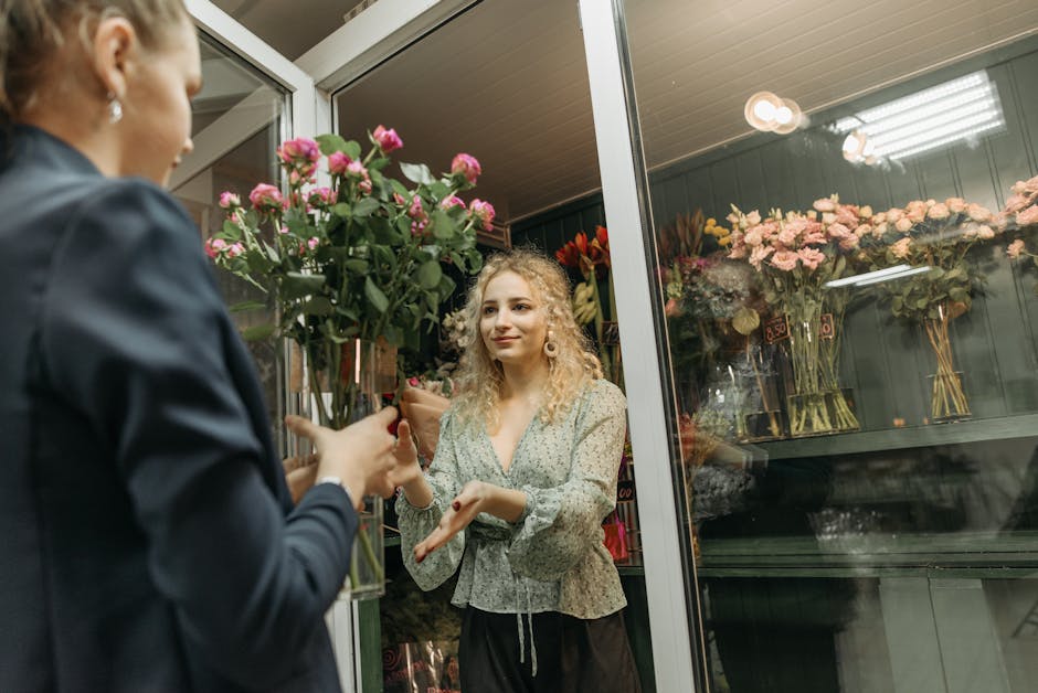 A woman with curly blonde hair, wearing a light-colored floral blouse, is seen inside a flower shop, receiving a bouquet of fresh pink roses with green foliage. The bouquet is wrapped in clear plastic