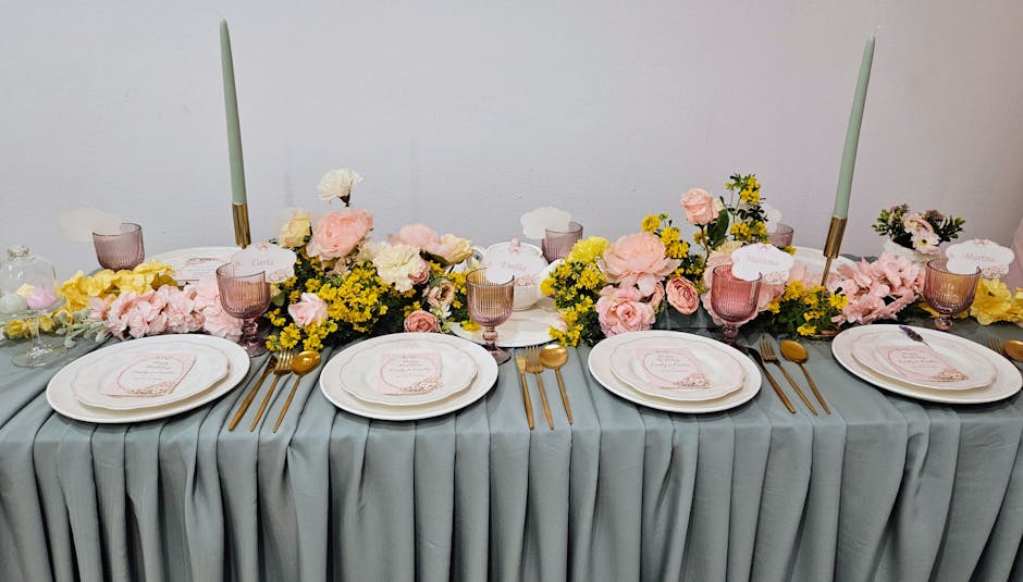 A floral arrangement featuring soft pink roses, yellow carnations, and delicate greenery displayed on a long table with a light grey, pleated tablecloth. The arrangement is interspersed with tall, sle