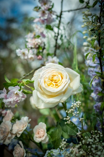 A close-up of a fresh, creamy white rose with softly layered petals, surrounded by delicate pink and white blossoms, and lush green foliage. The arrangement is set among various flowering stems and le