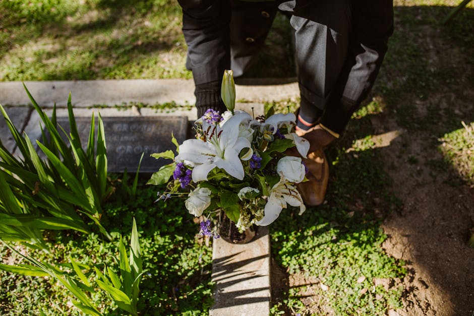 A person dressed in a dark overcoat, grey trousers, and brown shoes is placing a floral arrangement on a grave in an outdoor cemetery setting. The bouquet features prominent white lilies, with hints o