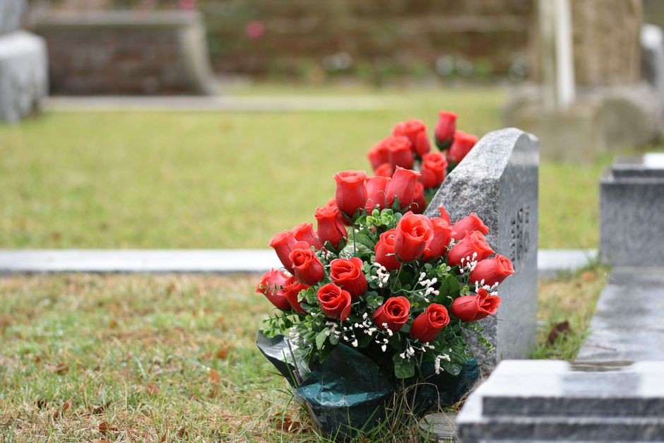 A bouquet of fresh red roses with tightly closed petals and green foliage, accented by small white filler flowers, arranged in a lush cluster and wrapped in dark green paper. The floral arrangement is